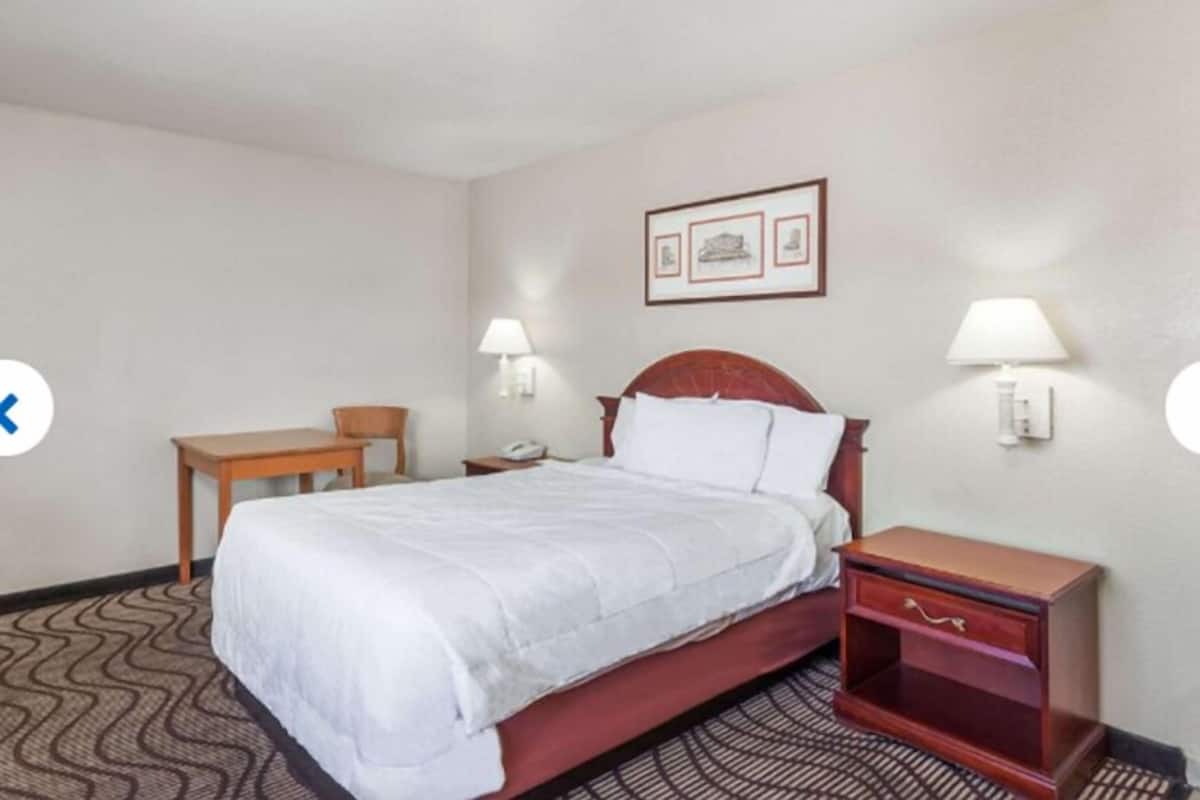 A hotel room with a single bed, a wooden nightstand, a desk, and two wall-mounted lamps.