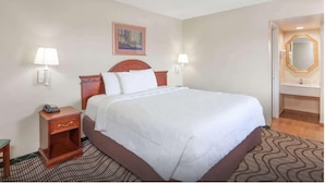 A hotel room with a large bed, bedside lamps, a small table with a phone, a painting on the wall, and a bathroom with a mirror and sink. - Tri City Inn (Victorville)