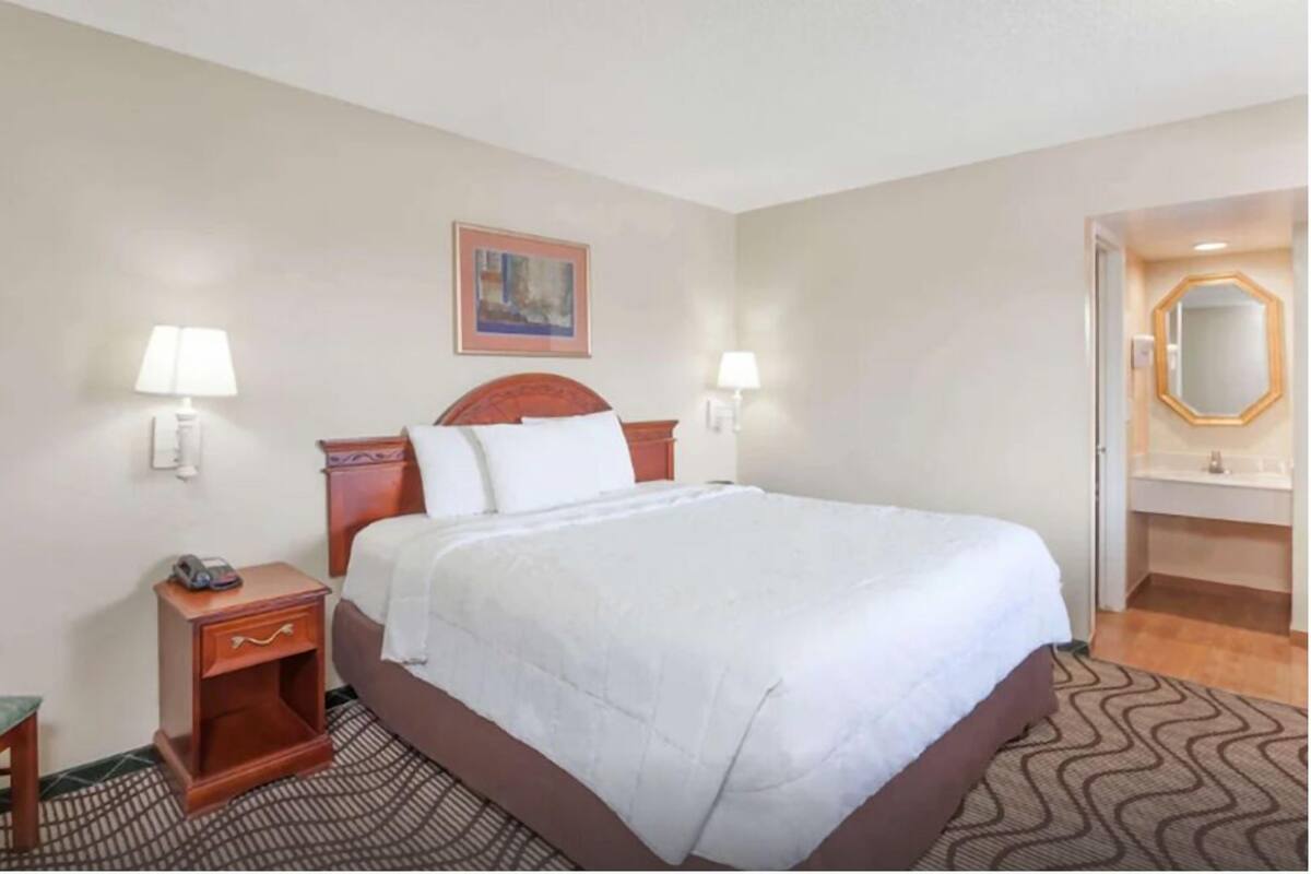 A hotel room with a large bed, bedside lamps, a small table with a phone, a painting on the wall, and a bathroom with a mirror and sink.