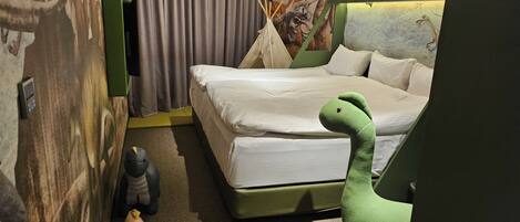 Une chambre d’hôtel avec un lit, un jouet en peluche vert, un petit jouet en forme d’éléphant et une fresque représentant un dinosaure sur le mur.