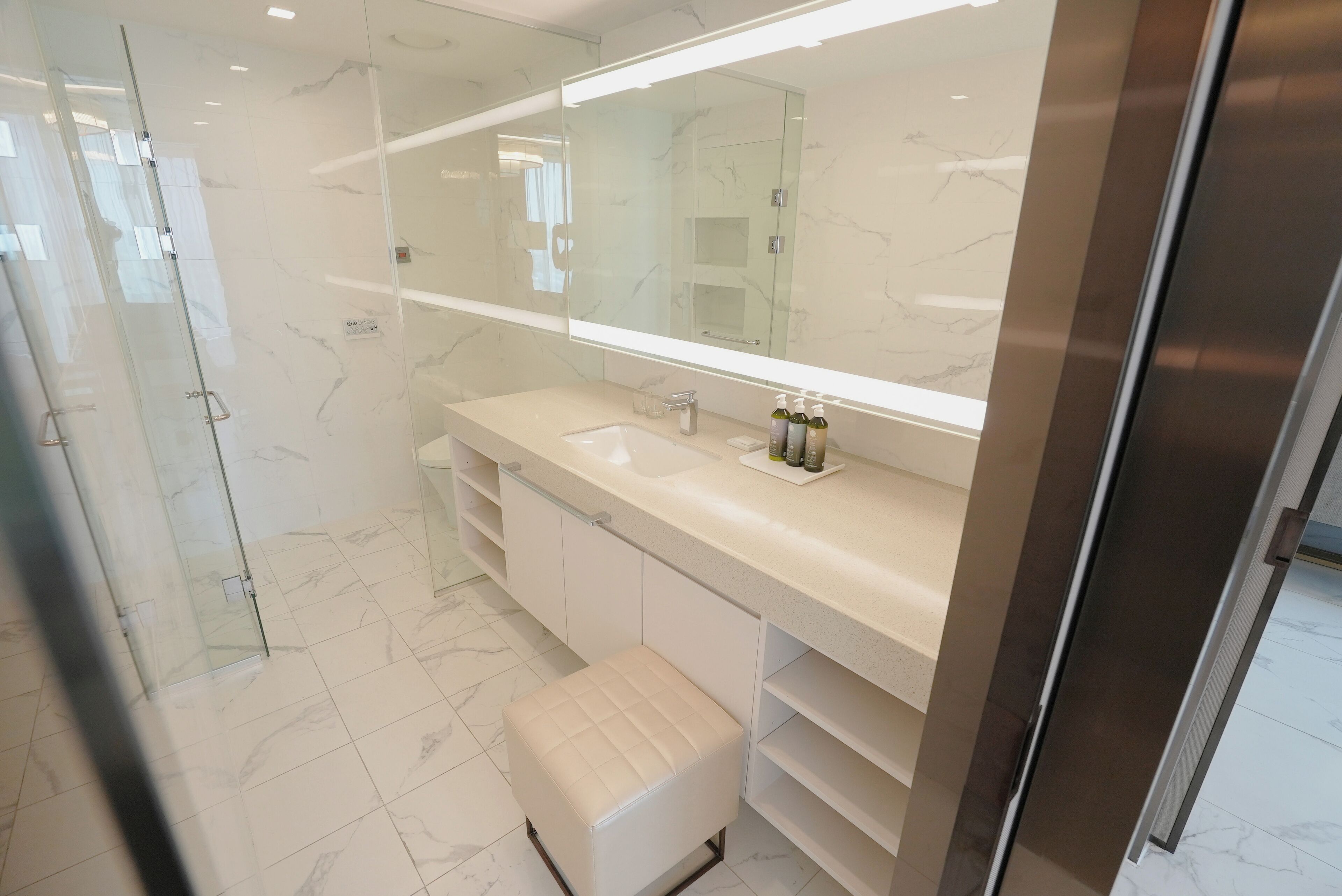 A modern bathroom with a large mirror, white marble tiles, a glass shower enclosure, and a beige bench.