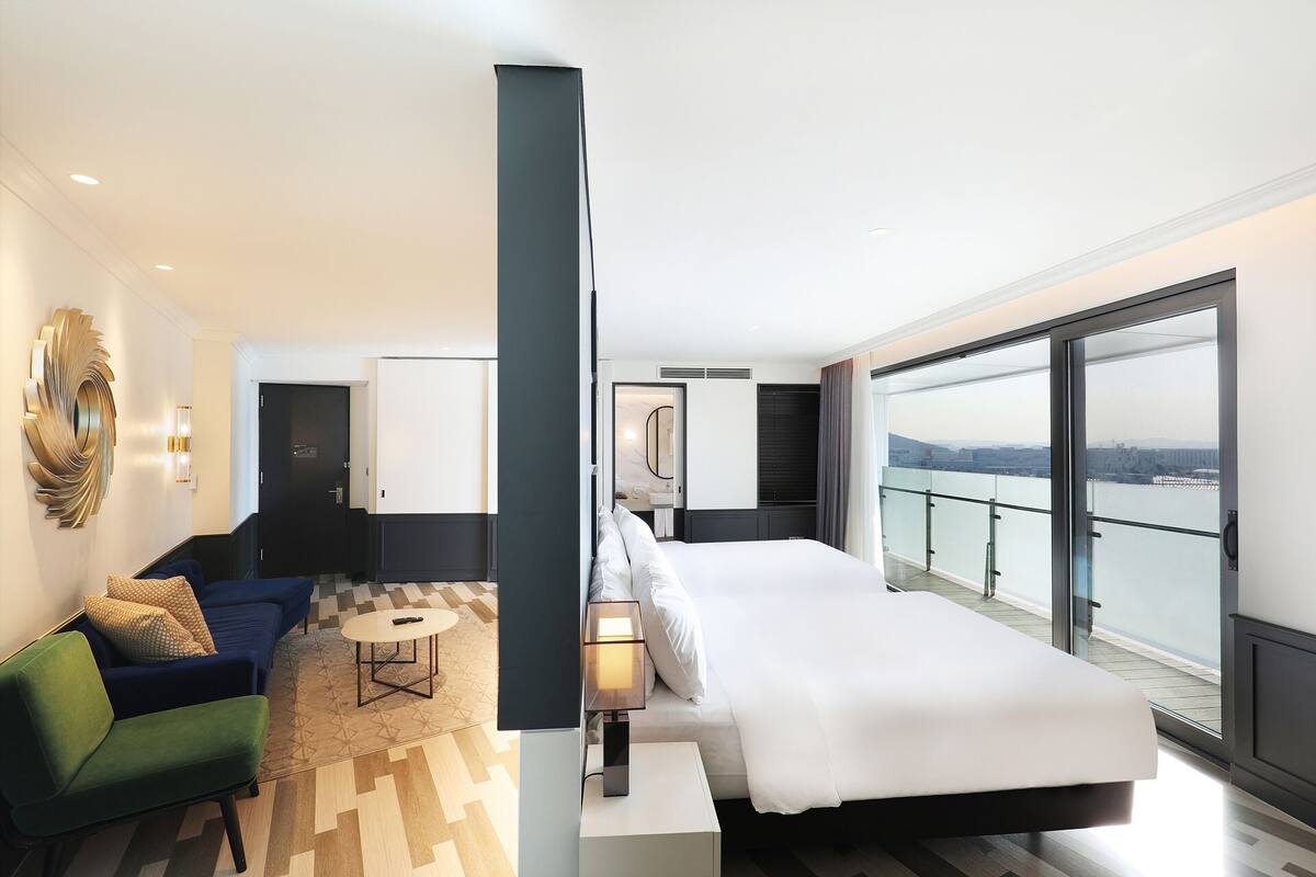 A modern hotel room with a large bed, a sofa, a small table, and a view of the city through large windows.