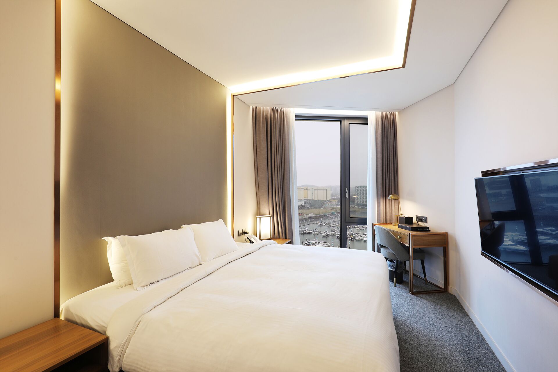 A modern hotel room with a large bed, a desk, a television, and a view of a marina through the window.