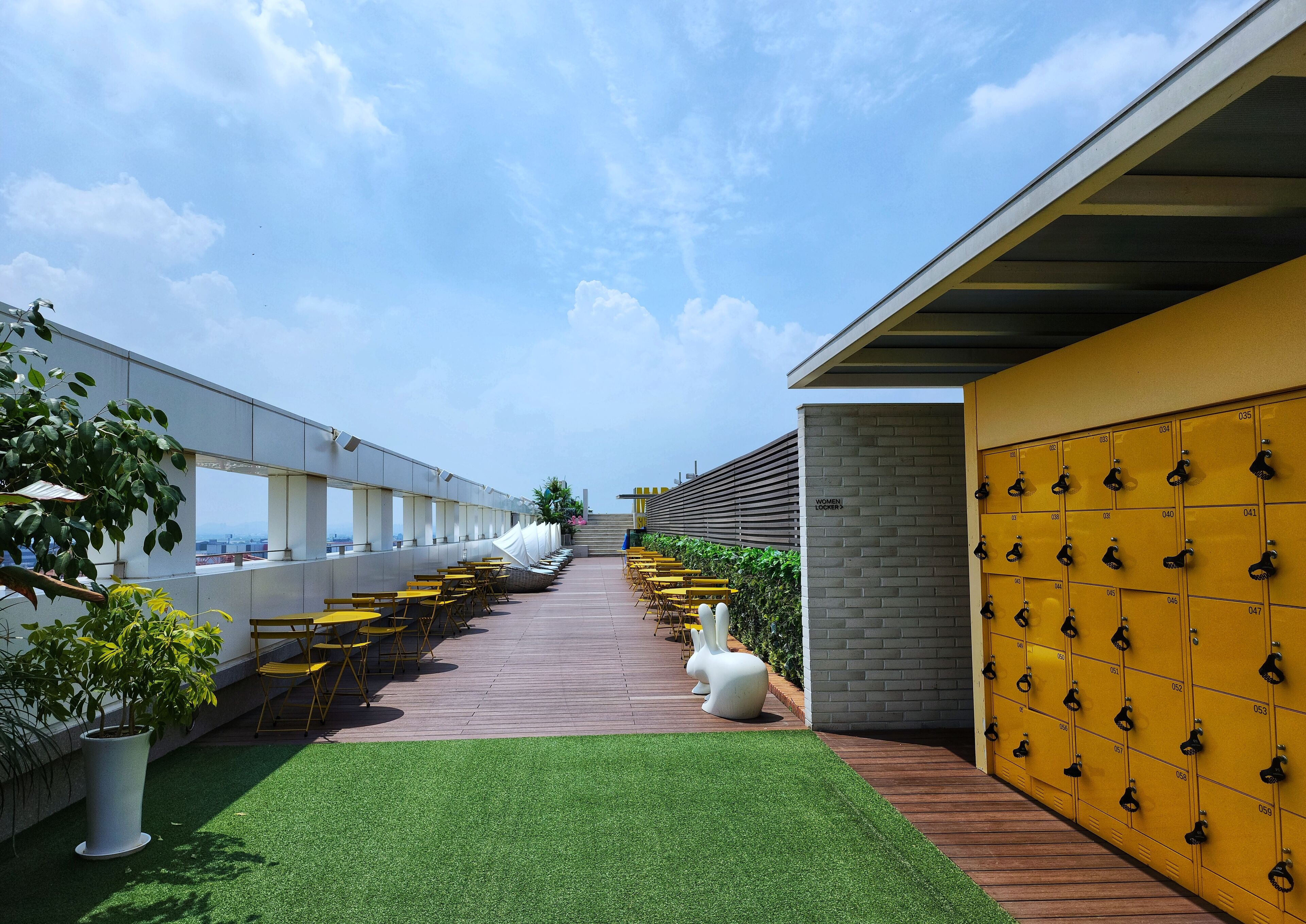 A rooftop terrace with yellow chairs and tables, a green lawn, and a row of yellow lockers.