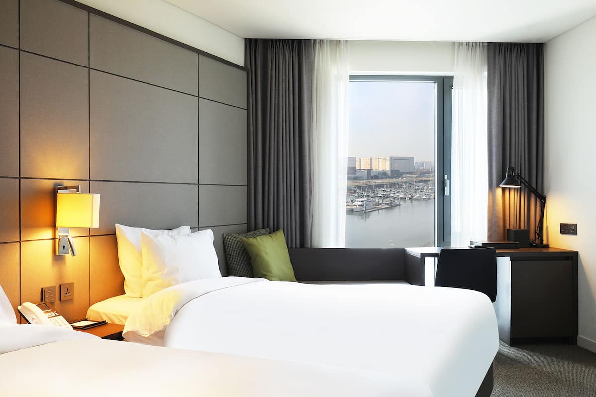 A hotel room with a large bed, a desk, a chair, a nightstand, a lamp, a phone, and a view of a marina and cityscape.