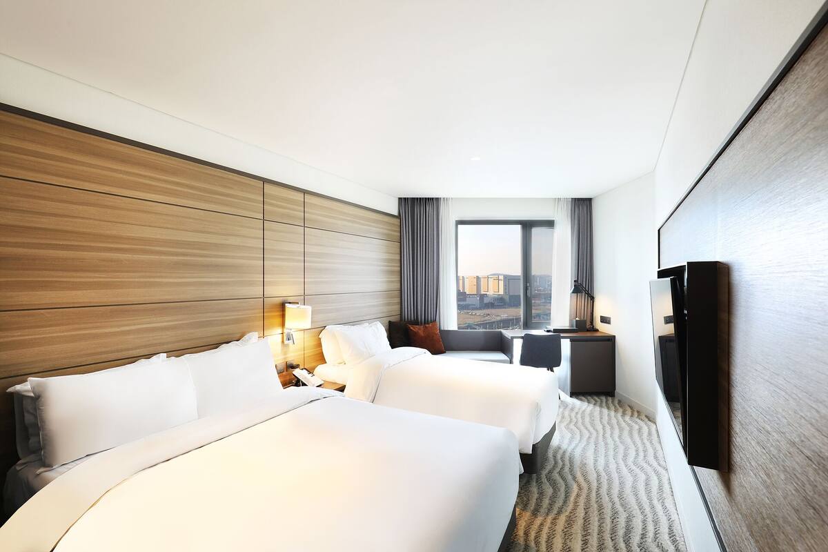 A hotel room with two beds, a desk, a TV, and a large window offering a city view.