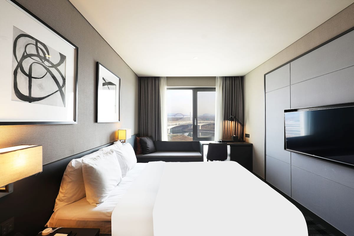 A hotel room with a large bed, a TV, a desk with a lamp, a chair, and a view of a bridge and landscape through the window.