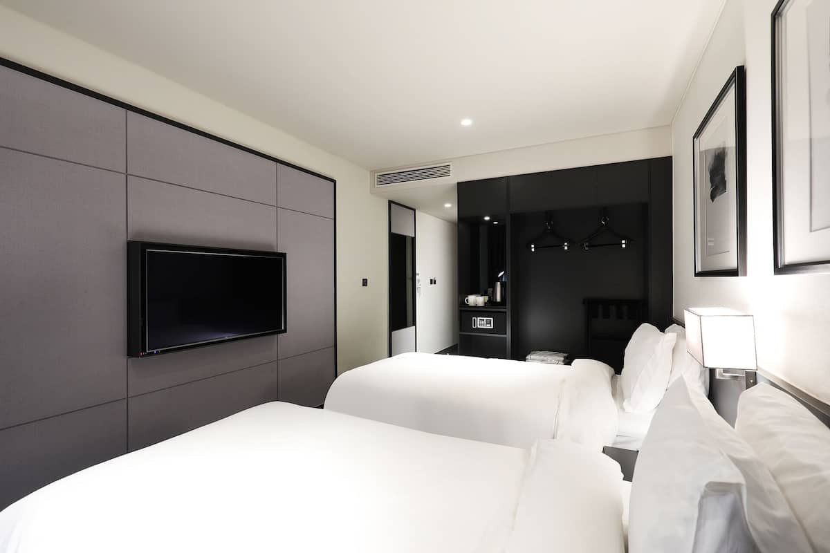 A modern hotel room with two beds, a flat-screen TV mounted on the wall, a bedside table with a lamp, and a mirror.