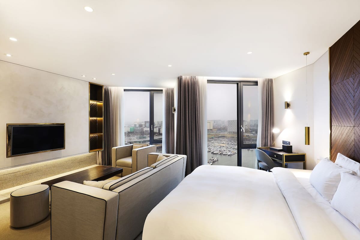 A modern hotel room with a large bed, a desk with a chair, a TV mounted on the wall, and a view of the cityscape through large windows.