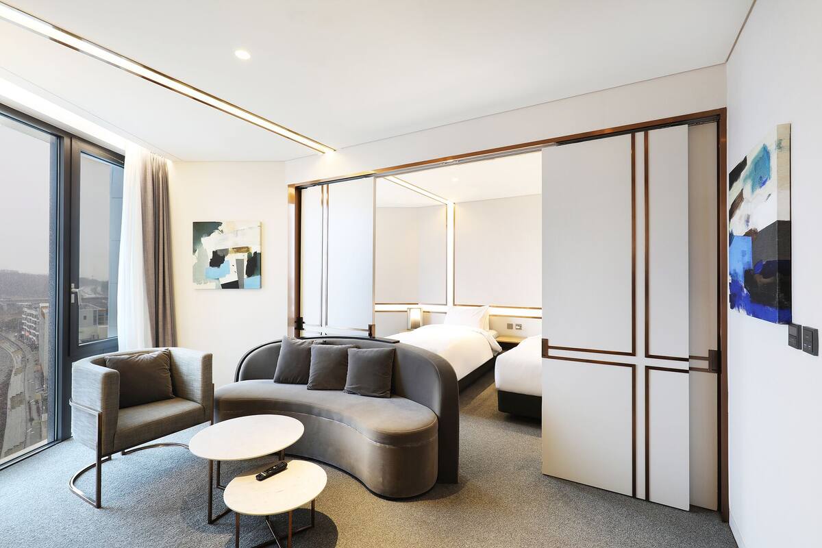 A modern hotel room with a sofa, two small tables, a bed, and a large window.