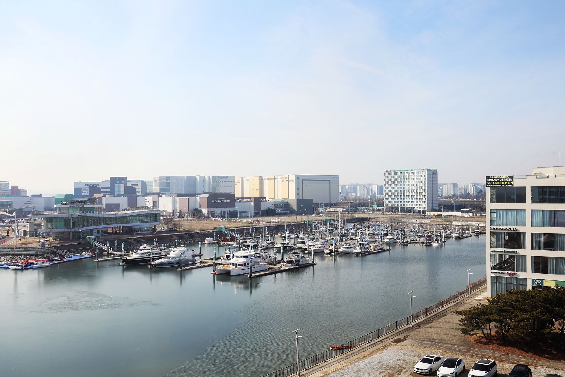 Un puerto deportivo con varias embarcaciones amarradas, un horizonte urbano al fondo y un edificio moderno con un letrero que dice "Premium Outlets".