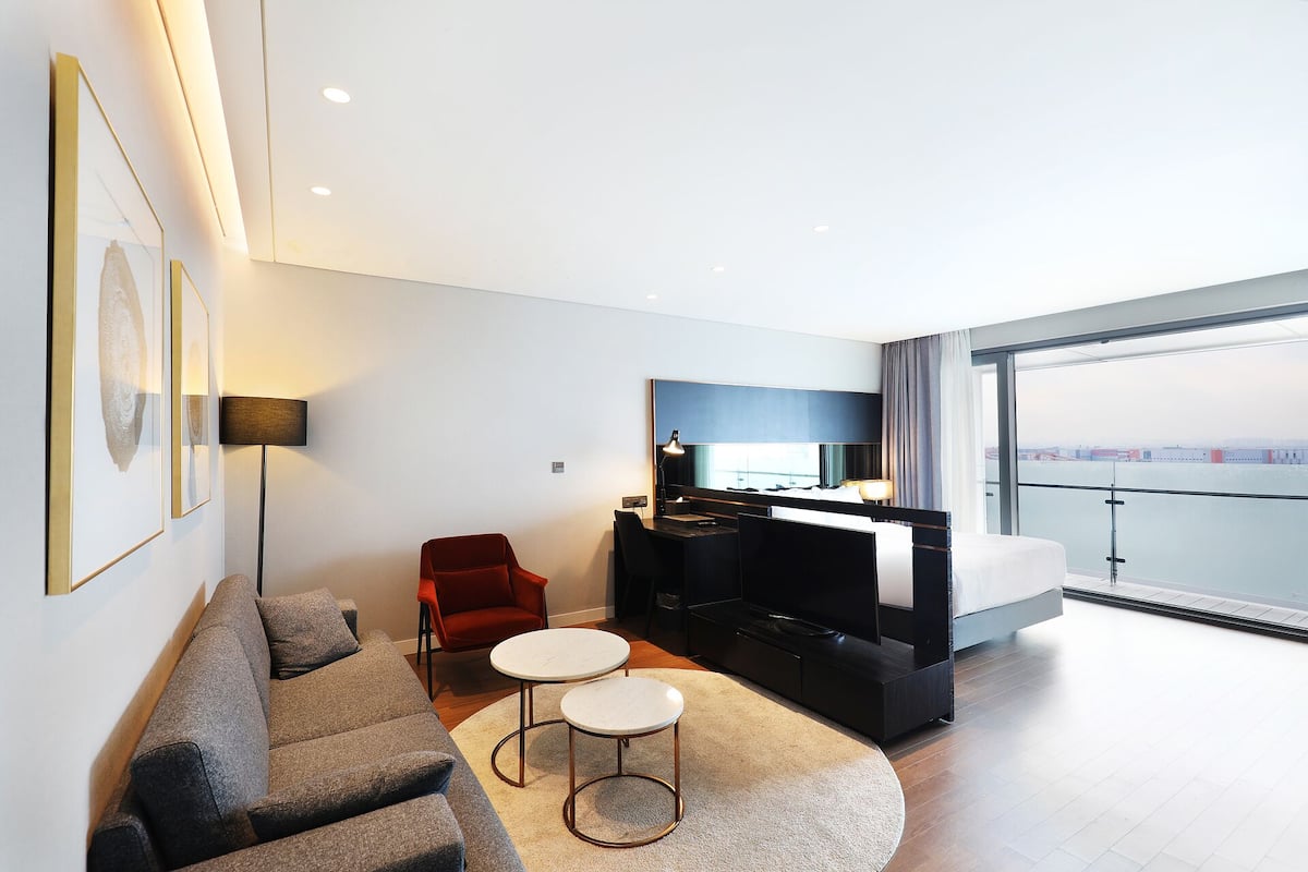 A modern hotel room with a bed, a desk, a red chair, a sofa, and a large window offering a city view.