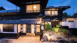 A traditional Japanese house with a tiled roof, wooden structure, and a small garden with rocks and a tree. - IZUTSURO (Tahara)