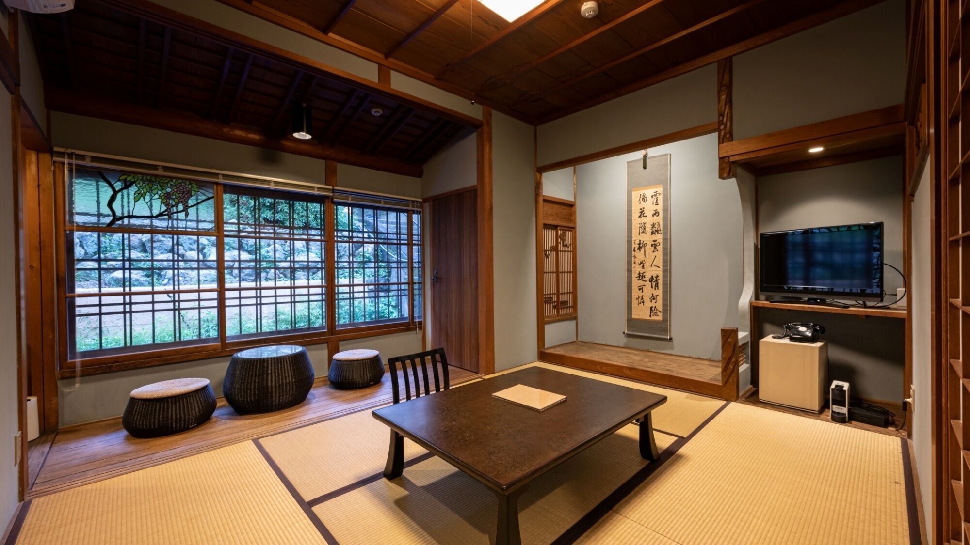 Ein traditioneller japanischer Raum mit Tatami-Boden, einem niedrigen Tisch, Kissen, einem Fernseher und einer Schiebetür mit Blick auf Grünflächen.