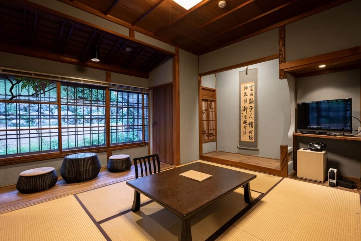 Ein traditioneller japanischer Raum mit Tatami-Boden, einem niedrigen Tisch, Kissen, einem Fernseher und einer Schiebetür mit Blick auf Grünflächen.