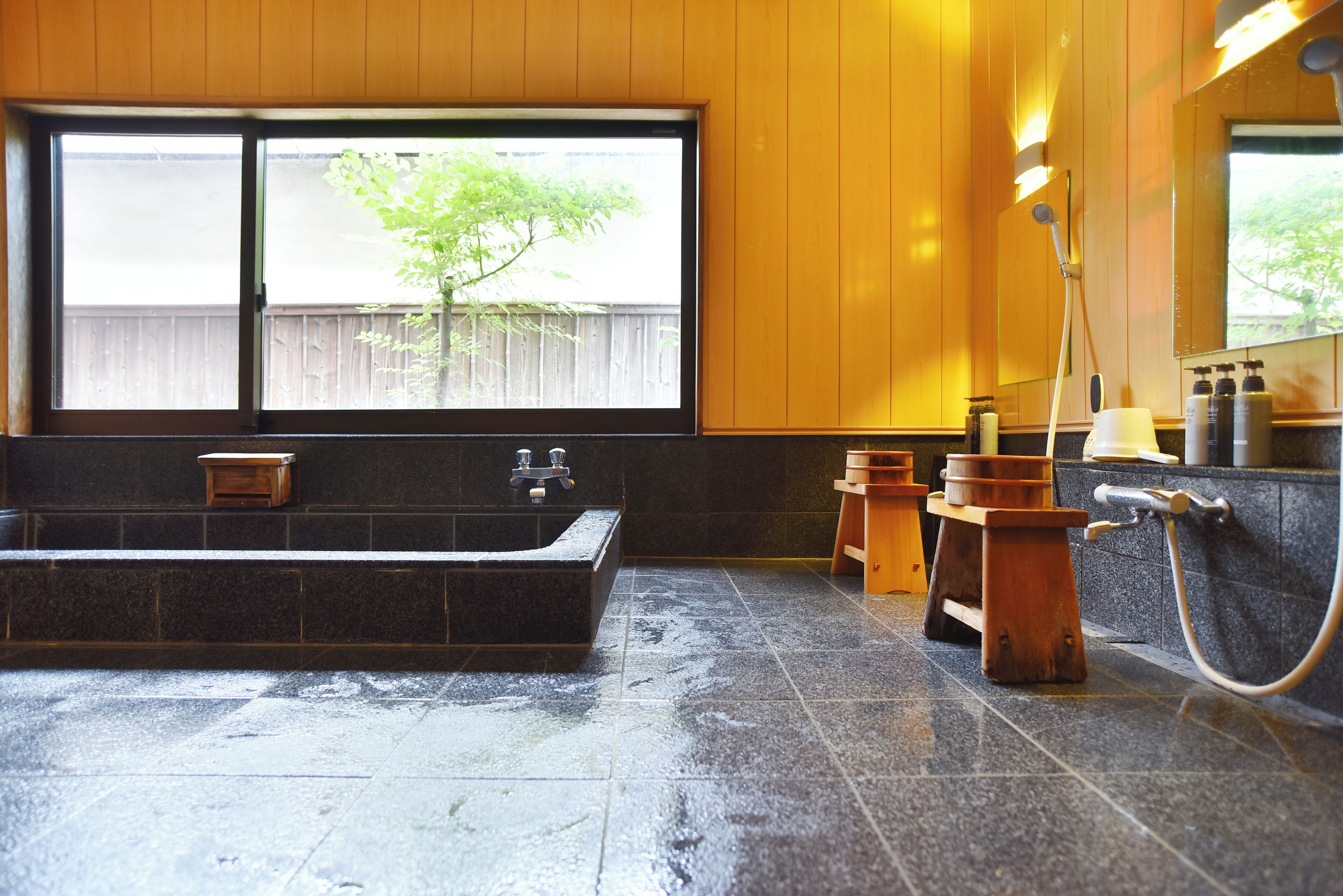 A bathroom with a large bathtub, wooden stools, a shower area, and a window with a view of a wooden fence and tree.