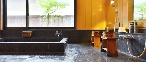 A bathroom with a large bathtub, wooden stools, a shower area, and a window with a view of a wooden fence and tree.