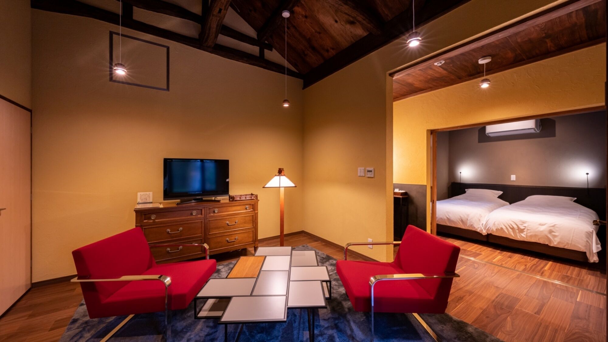 A hotel room with a wooden ceiling, a television, a lamp, a bed, and two red chairs.