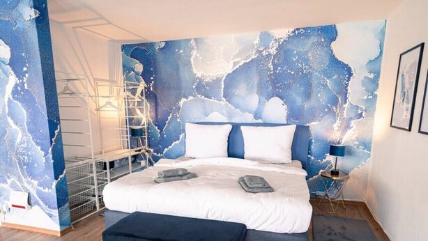 A modern bedroom with a large bed, a bench, a wall-mounted ladder, and a large abstract mural. - City Loft | 5 Min Rhein-Galerie (Ludwigshafen am Rhein)