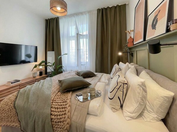 A modern hotel room with a large bed, a flat-screen TV, a bedside table with a lamp, and a window with curtains.