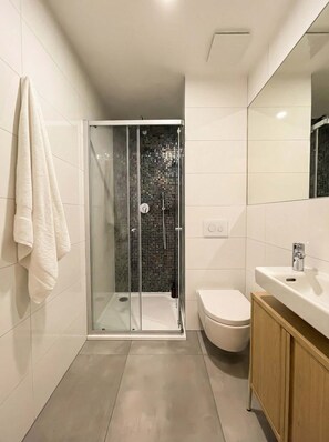 A modern bathroom with a glass shower enclosure, a white toilet, and a wooden vanity. - TurnKey | Angelo Roma Studio V (Praha 2)
