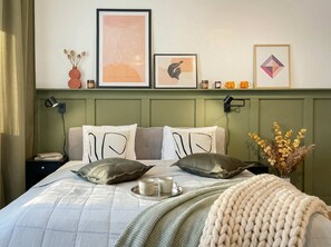 Un dormitorio ordenado con cama, almohadas, una mesita de noche, una lámpara y decoración en la pared.