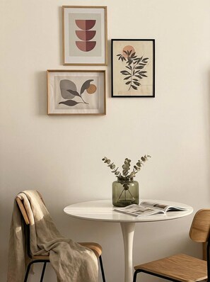 Una habitación con una mesa redonda, una silla, un pequeño jarrón con flores secas y tres obras de arte enmarcadas en la pared.