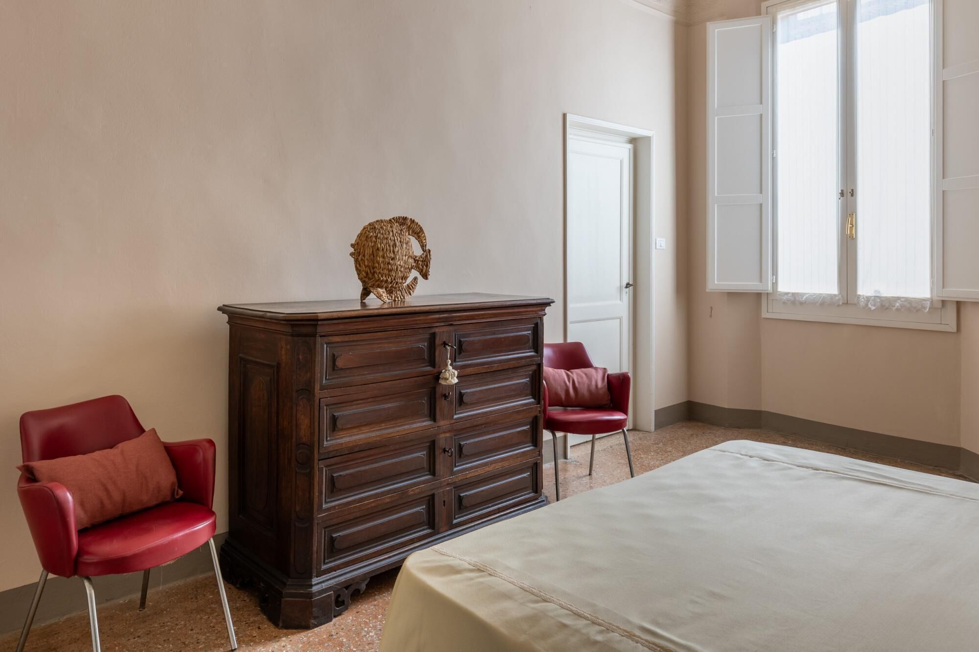 Une chambre à coucher avec une commode en bois, deux chaises rouges, un lit et une fenêtre avec des rideaux blancs.