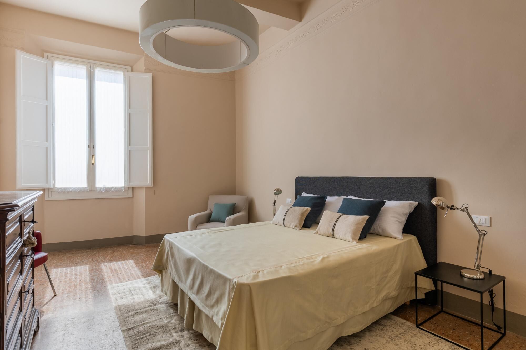 Ein Schlafzimmer mit Bett, Nachttisch, Lampe, Stuhl und einem Fenster mit Vorhängen.