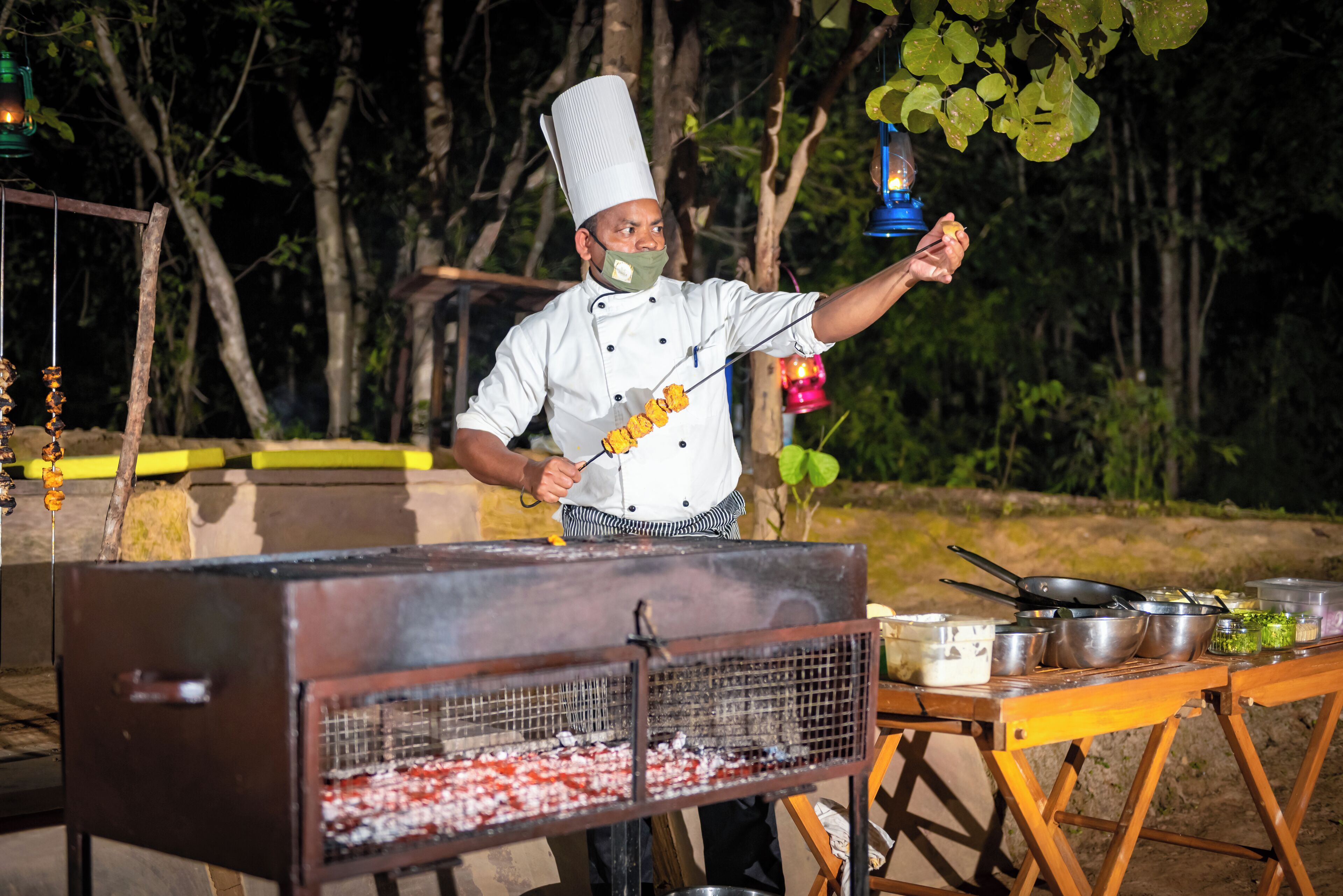 Un chef asando al aire libre con una parrilla de carbón, utensilios de cocina y una mesa plegable con equipo culinario.