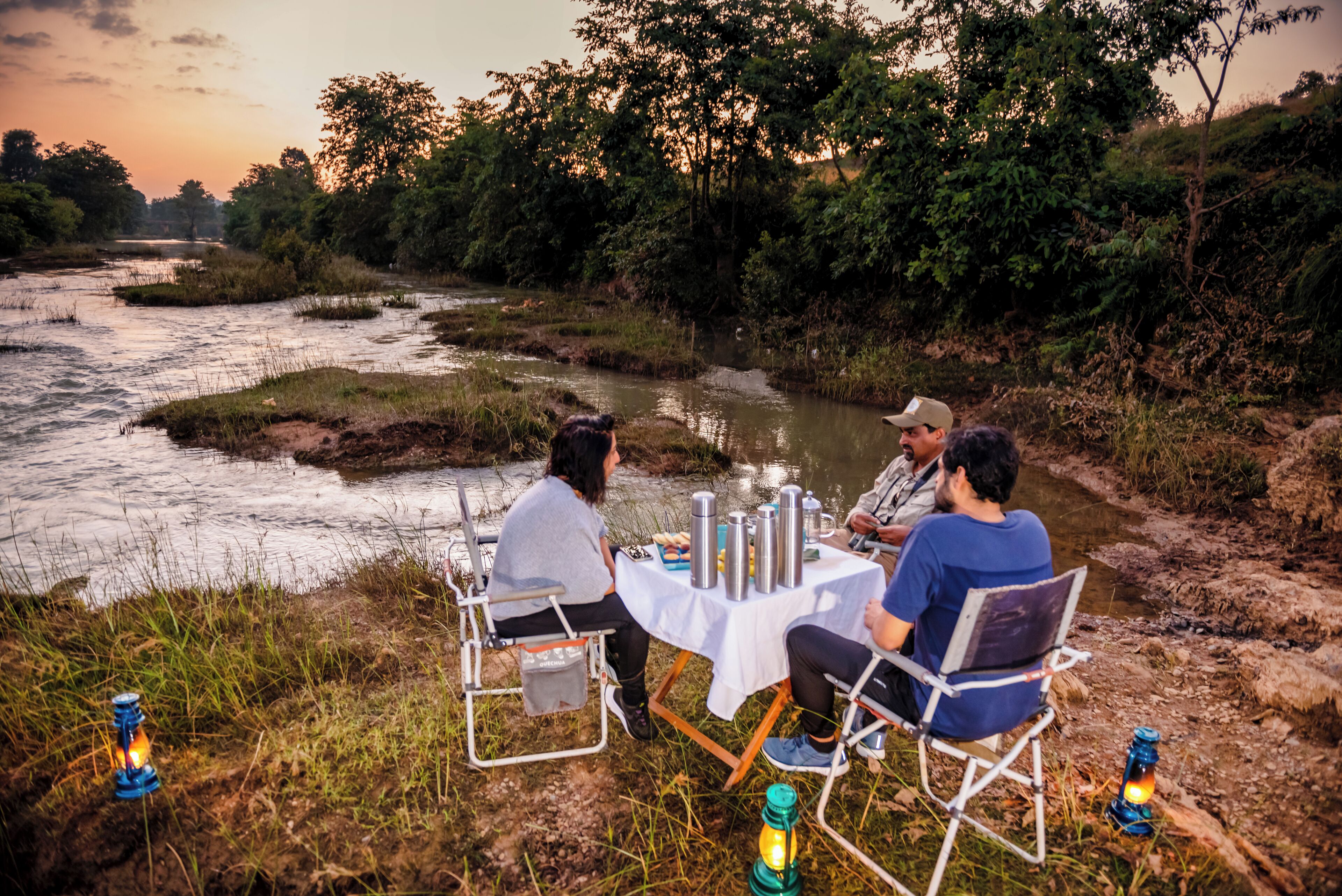 Drie personen zitten aan een tafel met campingstoelen, een tafelkleed en staan er kampverlichtingen op de grond.