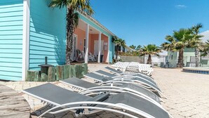 A row of sun loungers aligned along a wooden deck, a turquoise building with a porch, and palm trees in the background. - 6007 Rookery 3 Just Breathe (Gulf Shores)