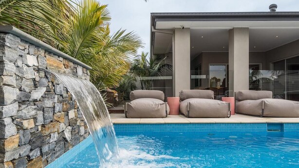 Una piscina con cascada de piedra, sillas reclinables color beige y una casa moderna al fondo.