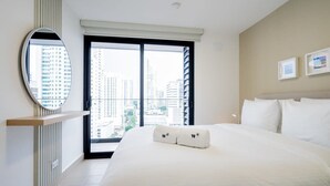 A hotel room with a large bed, a round mirror, a window with a view of tall buildings, and two framed pictures on the wall.