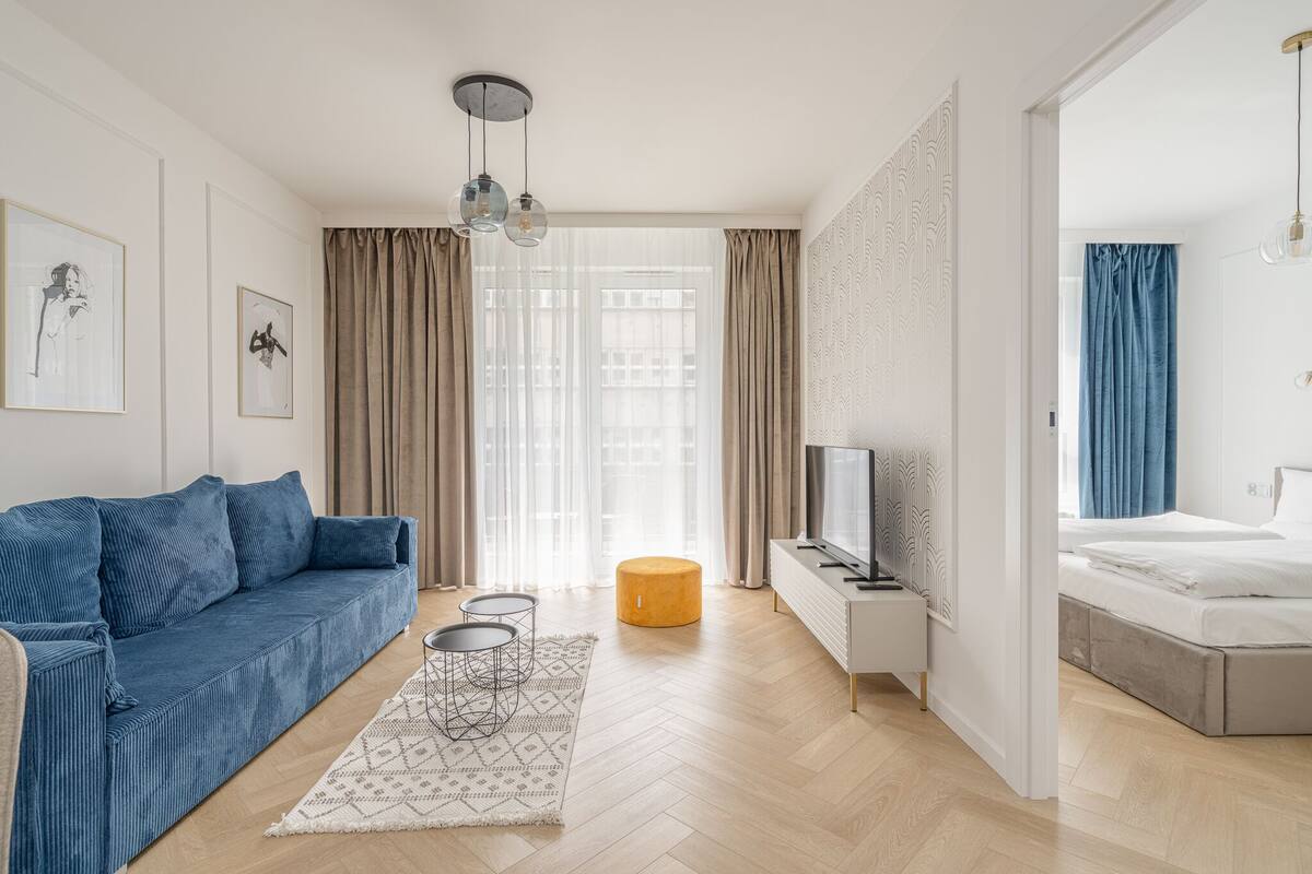 A modern living room with a blue sofa, a small round ottoman, a TV, and a bed in the adjacent room.