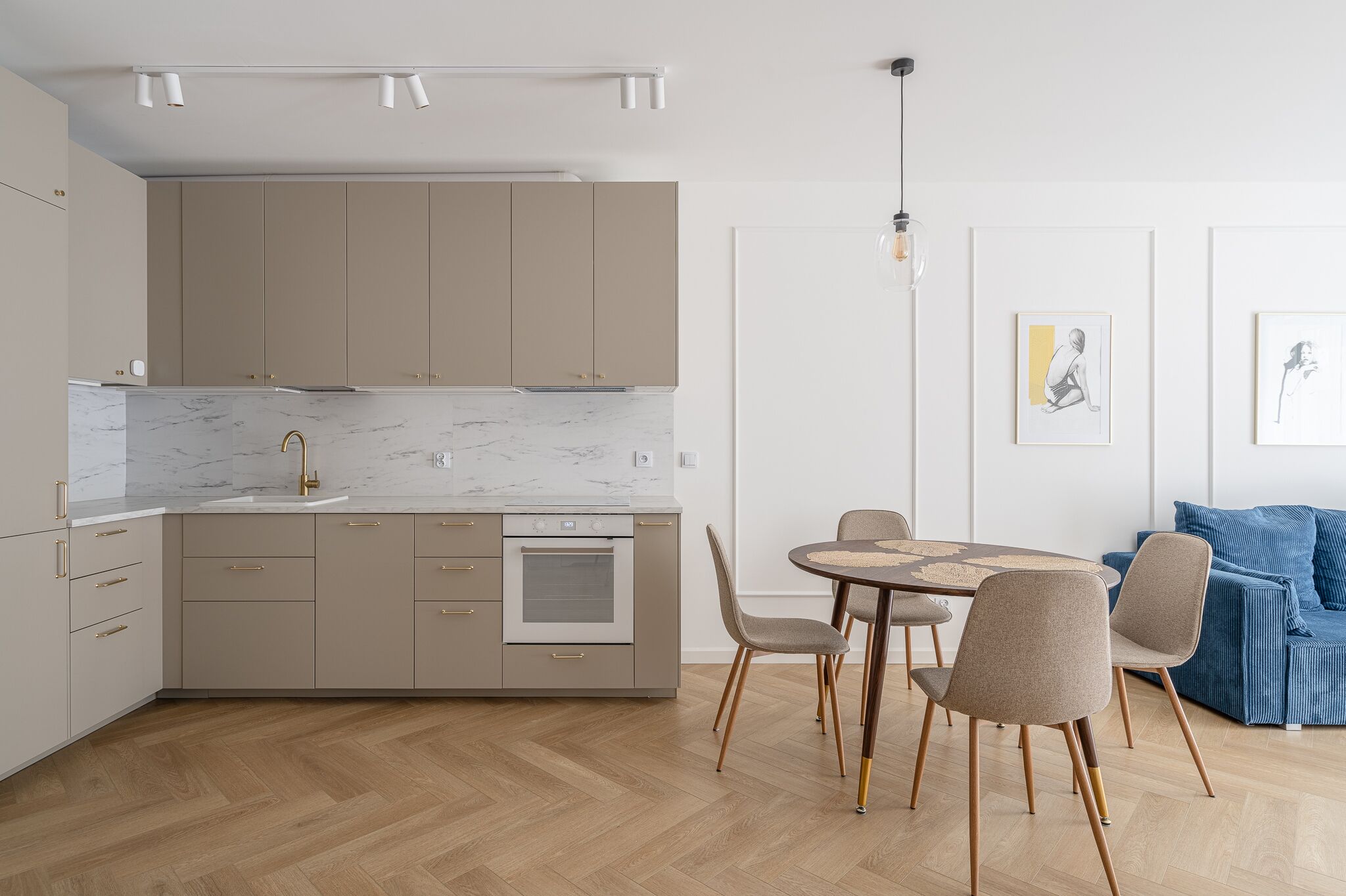 A modern kitchen with beige cabinets, a marble countertop, and a built-in oven. There is a dining area with a round table and chairs.