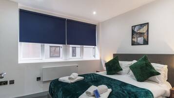 A hotel room with a bed, a window with blinds, a radiator, and a painting on the wall.