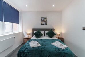 A hotel room with a bed, two pillows, a nightstand, a lamp, a window with a blind, and a painting on the wall.