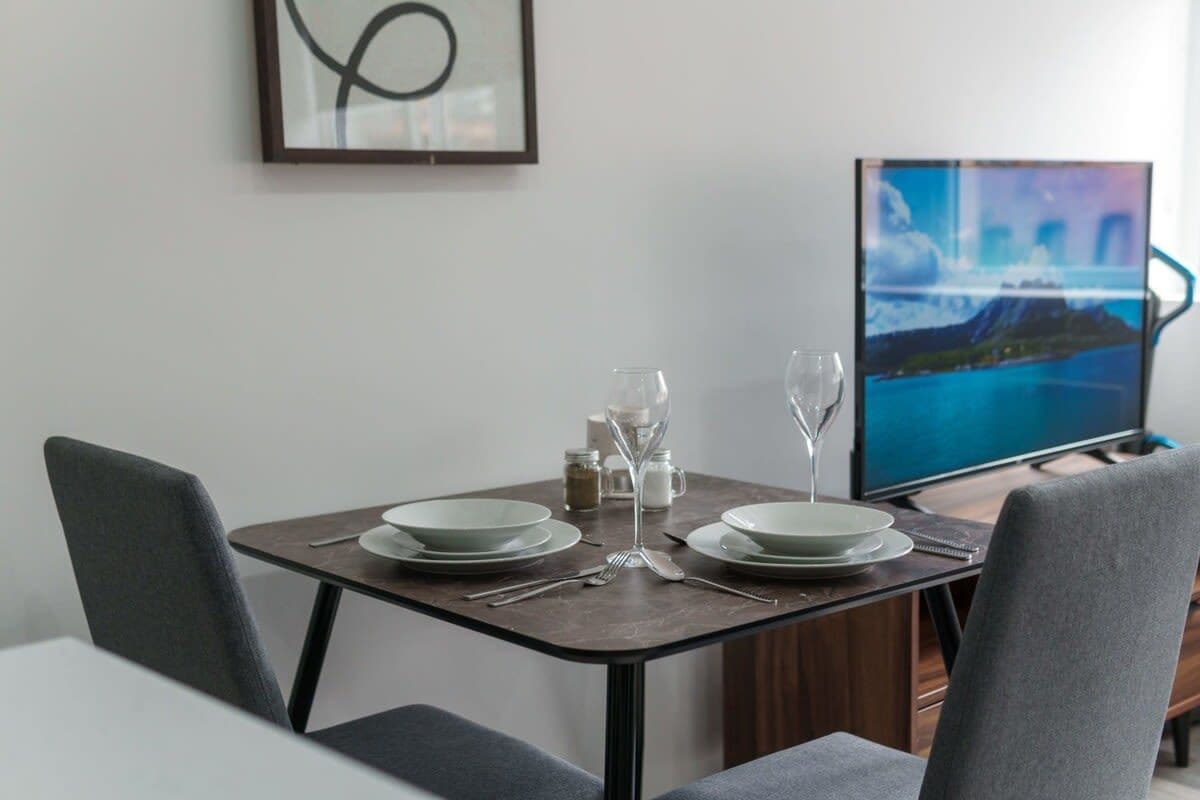 A dining area with a table set for two, a television displaying a scenic landscape, and a framed abstract artwork on the wall.