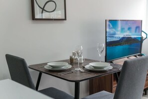 A dining area with a table set for two, a television displaying a scenic landscape, and a framed abstract artwork on the wall. - Central Preston Contemporary 1 Bedroom Apartment (Preston)