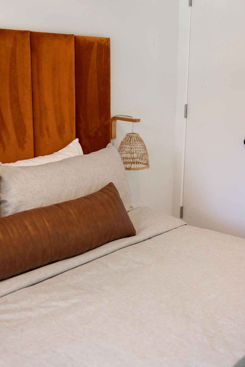 A neatly made bed with a wooden headboard, a woven lamp, and a white door.