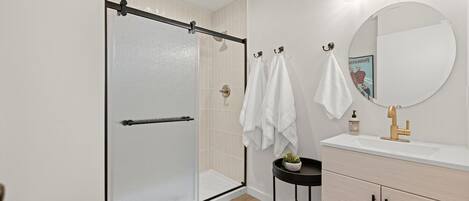A modern bathroom with a glass shower enclosure, a round mirror, a sink with a gold faucet, and a small round table with a plant.
