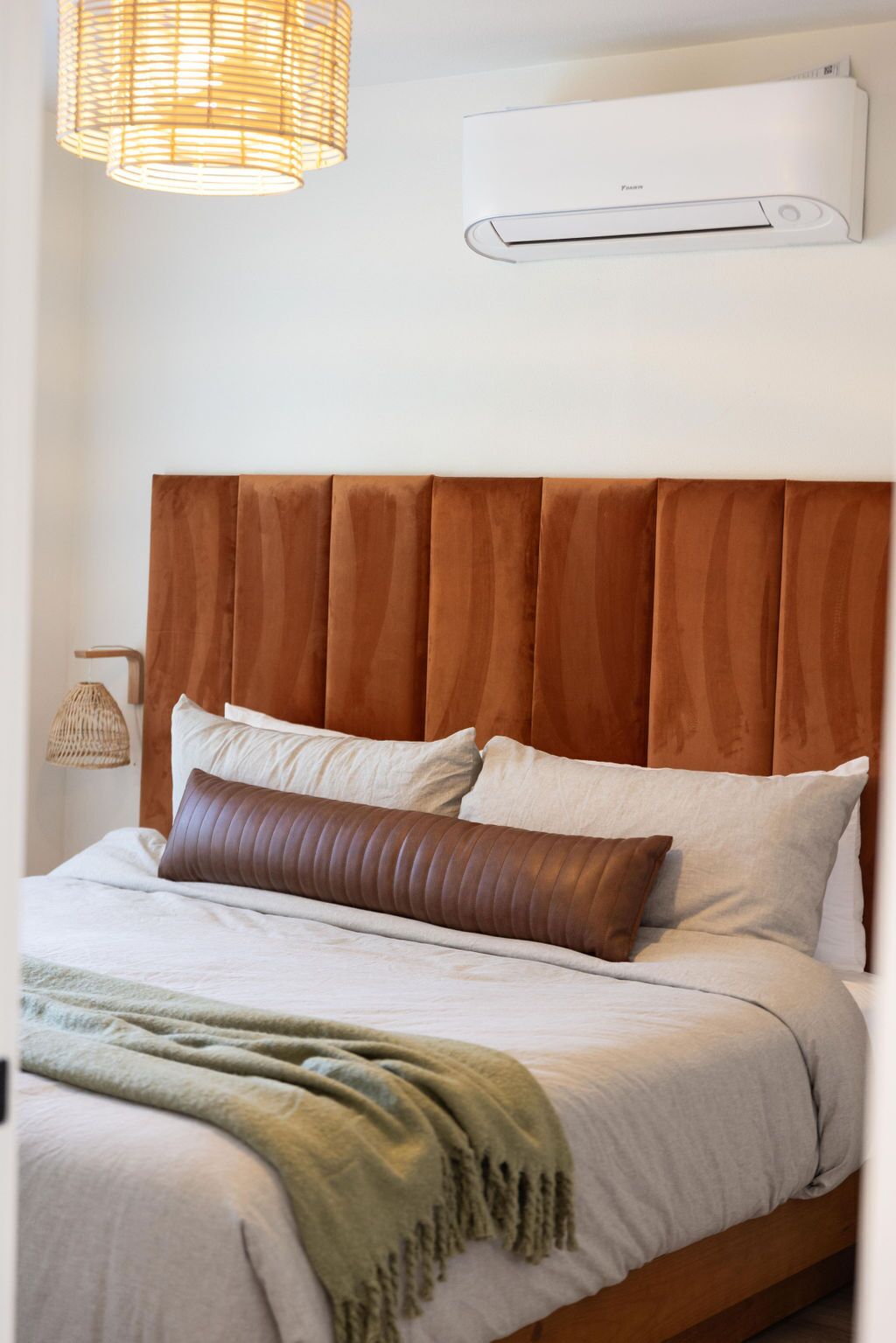 Ein ordentlich bezogenes Bett mit Holz-Kopfteil, einer gewebten Lampe und einer Wand-Klimaanlage.