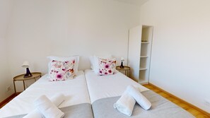 A double bed with floral pillows, a bedside table with a lamp, a wardrobe, and a rolled towel on the bed.