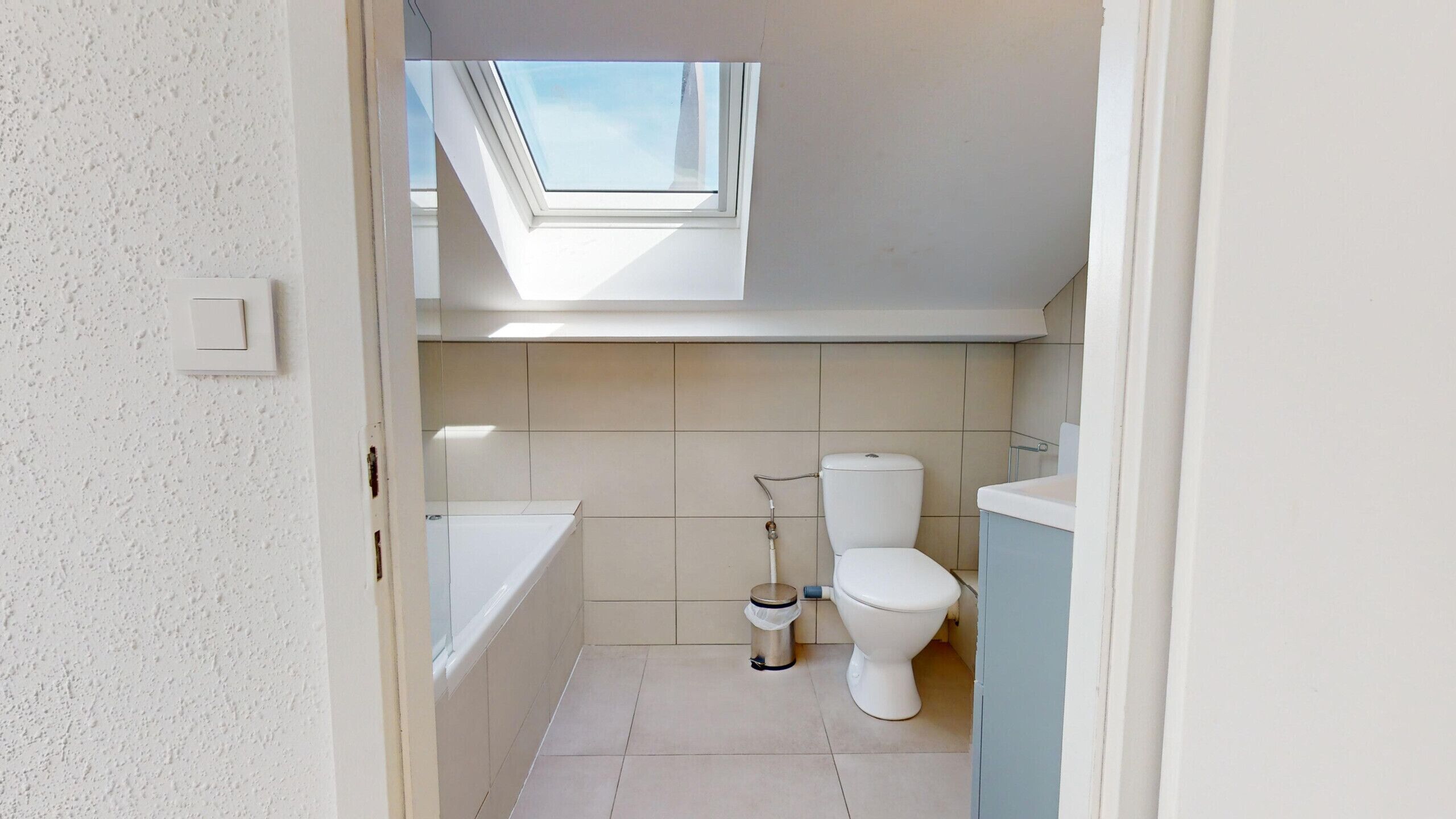 Un baño pequeño y moderno con tragaluz, inodoro, bañera y un cubo de basura.