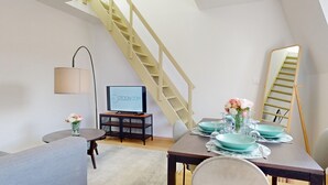 A modern living room with a staircase, a television, a dining table set with plates and glasses, and a vase with flowers.