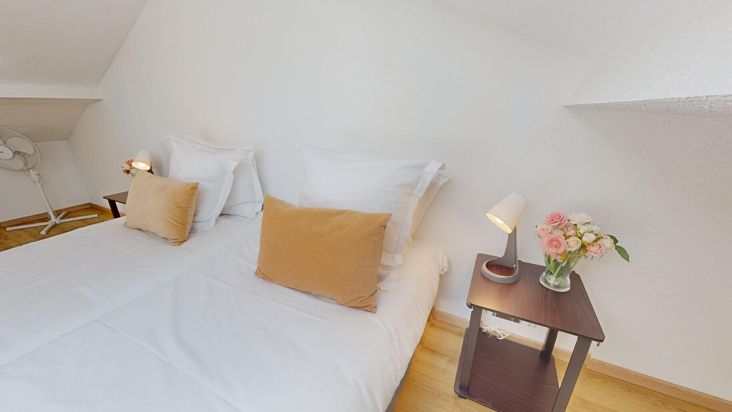 Een slaapkamer met een bed, kussens, een nachtkastje met een lamp en een vaas met bloemen.