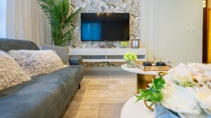 A living room with a grey sofa, a flat-screen TV mounted on the wall, a large potted plant, and a small round table with decorative items. - Majestic & Spacious1b Downton Gym Pool (Santo Domingo)