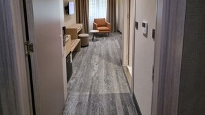 Un couloir d’hôtel avec un sol recouvert de moquette, un coin salon comprenant une chaise et un pouf, une petite table et un téléviseur fixé au mur.