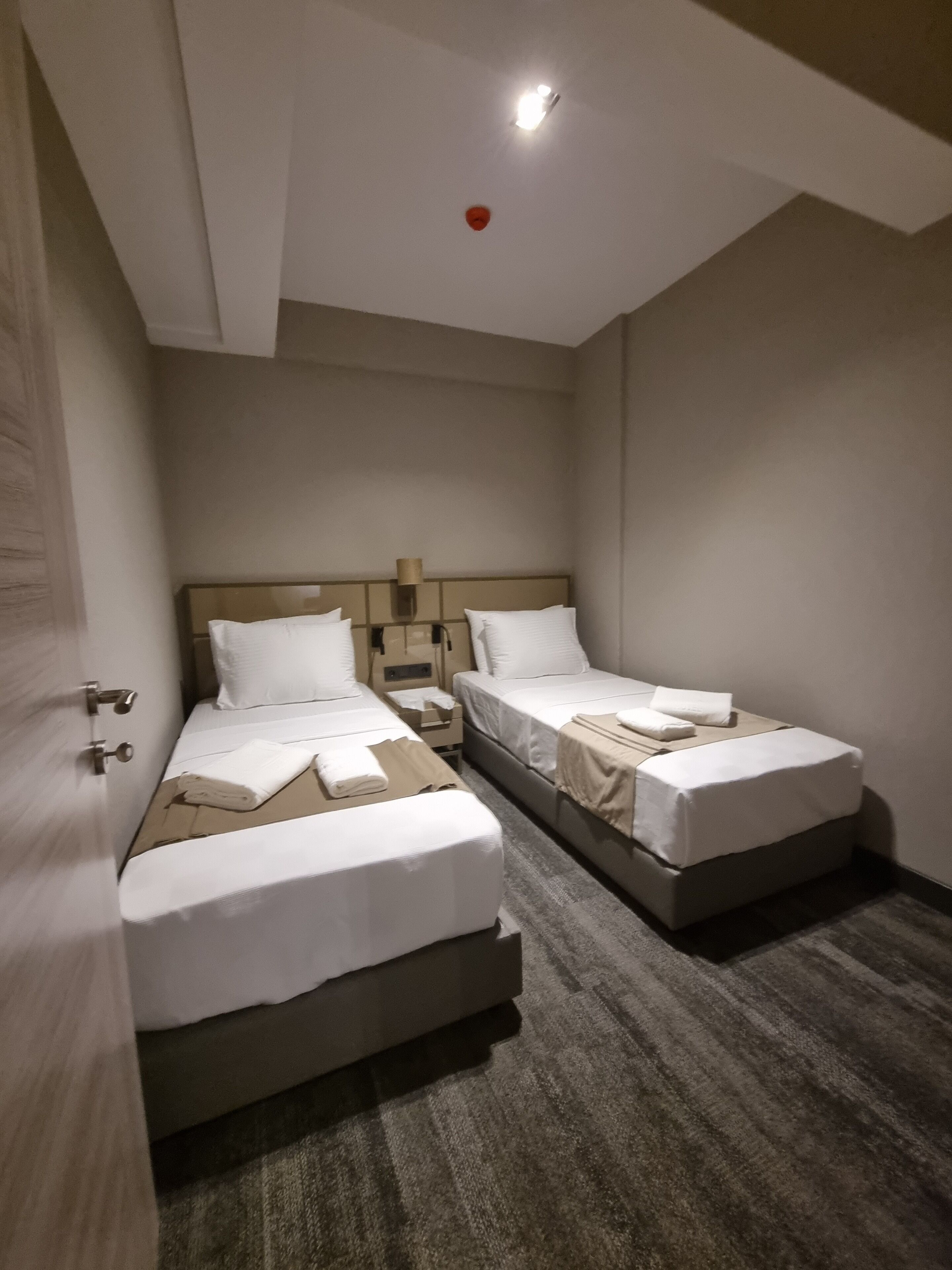 A hotel room with two single beds, a nightstand, a lamp, and a red emergency light on the ceiling.